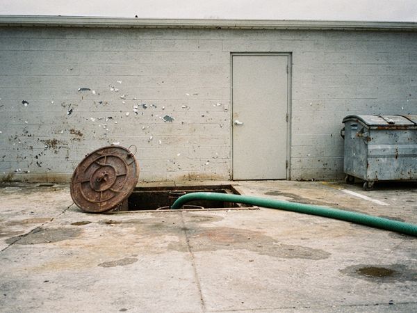 An open grease trap in the concrete behind a restaurant, its round cover set aside and a green vacuum hose running into it.