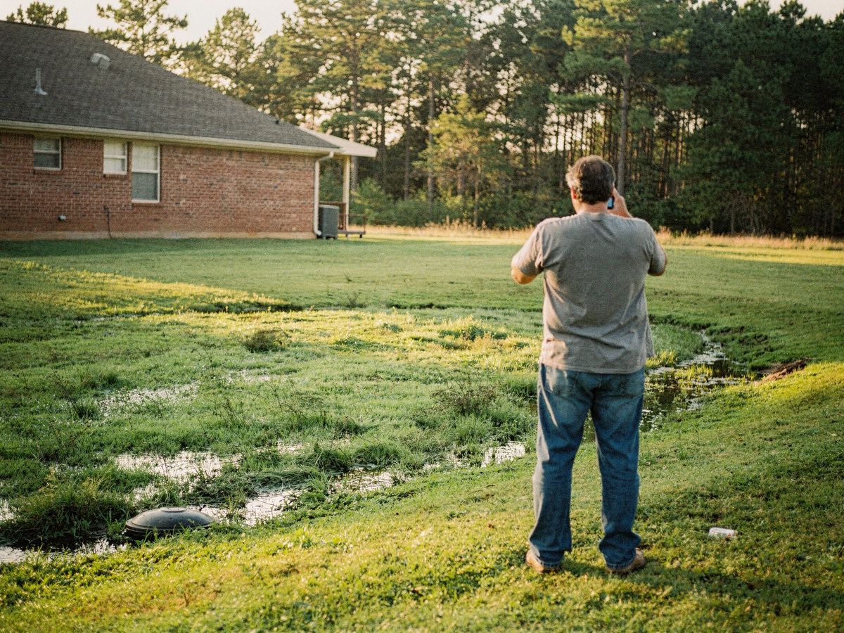 A homeowner, seen from behind, on the phone while looking at a soggy, waterlogged patch of back yard.