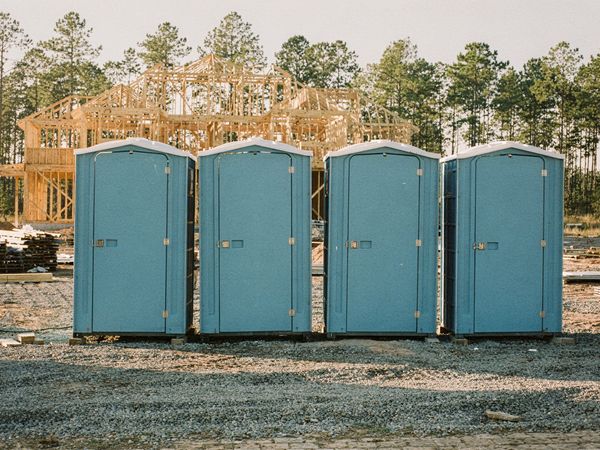 Four plain blue portable toilets on gravel at a house construction site, with pine trees behind.