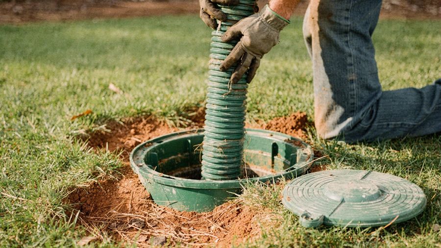 Gloved hands feeding a green suction hose into an open septic tank riser in a grassy yard.