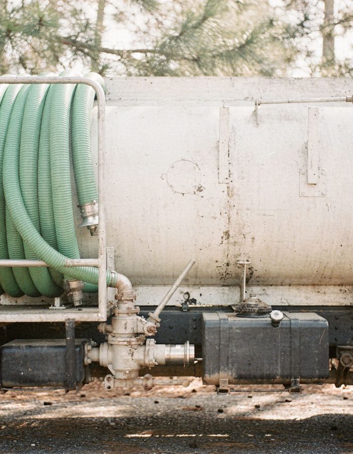 The side of a plain white vacuum pump truck with a coiled green suction hose on its rack, parked under pine trees.